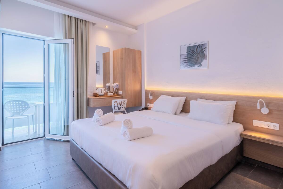 A hotel room with a large bed, a desk with a chair, a view of the sea, and a balcony with a chair.