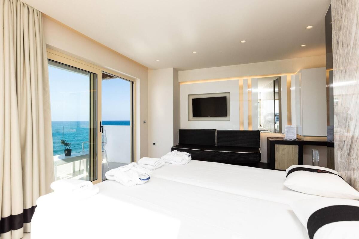 A hotel room with a large bed, a TV mounted on the wall, and a balcony with a view of the sea.
