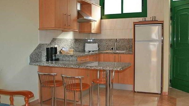 Eine kompakte Küche mit Holdekor-Schränken, einer Granitarbeitsplatte, einem eingebauten Backofen und einem Kühlschrank. Es sind drei Barhocker und ein Holenstuhl vorhanden.