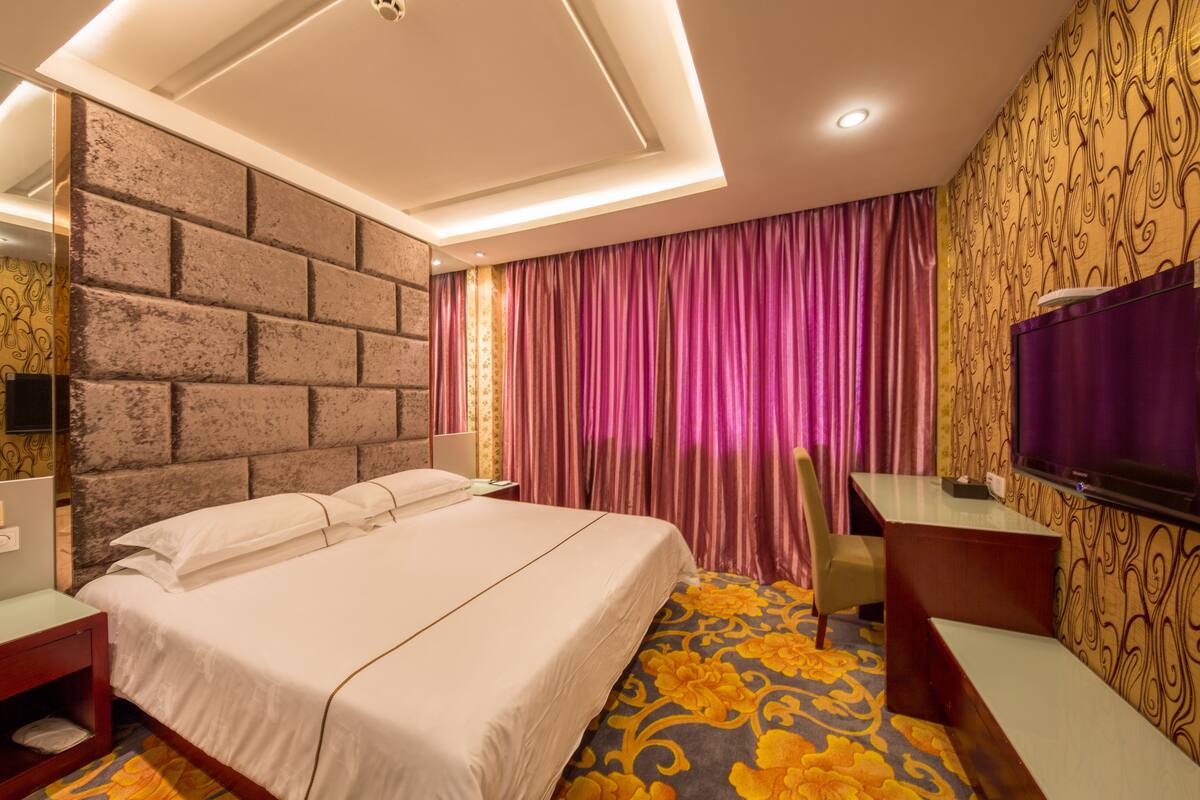 A hotel room with a large bed, a desk, a TV, and a patterned carpet.