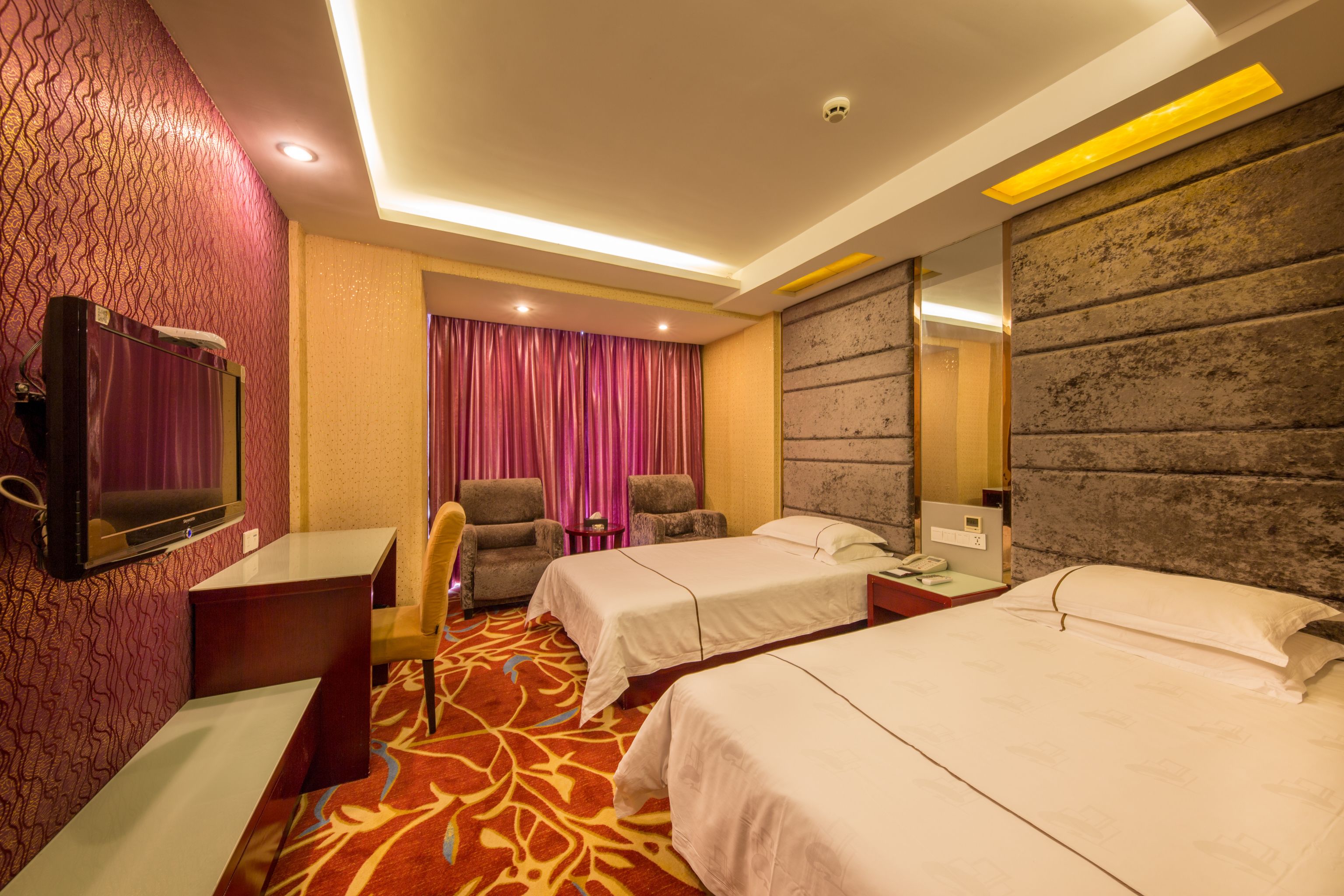 A hotel room with two beds, a desk, a chair, a television, and a patterned carpet.