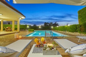 Zona junto a la piscina con sillones de descanso, una mesa con bebidas y vista al agua.