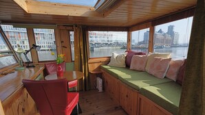 A cabin with a wooden interior, a dining area with a table and chairs, a window seat with cushions, and a view of a cityscape and water. - Cozy and romantic apartment in captain's steering cabin (Amsterdam)