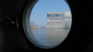 A view through a circular window of a modern building across a body of water. - Cozy and romantic apartment in captain's steering cabin (Amsterdam)