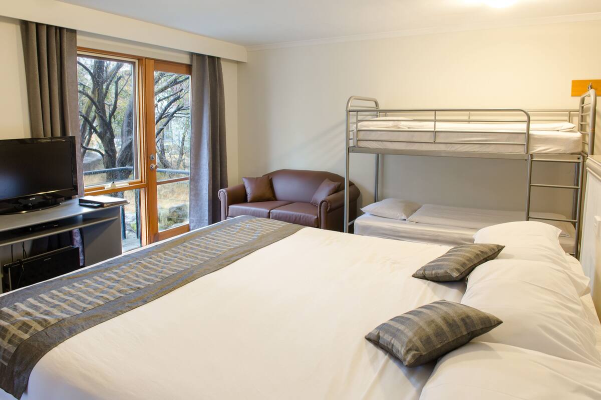 A hotel room with a large bed, a TV on a desk, a bunk bed, and a window with curtains.