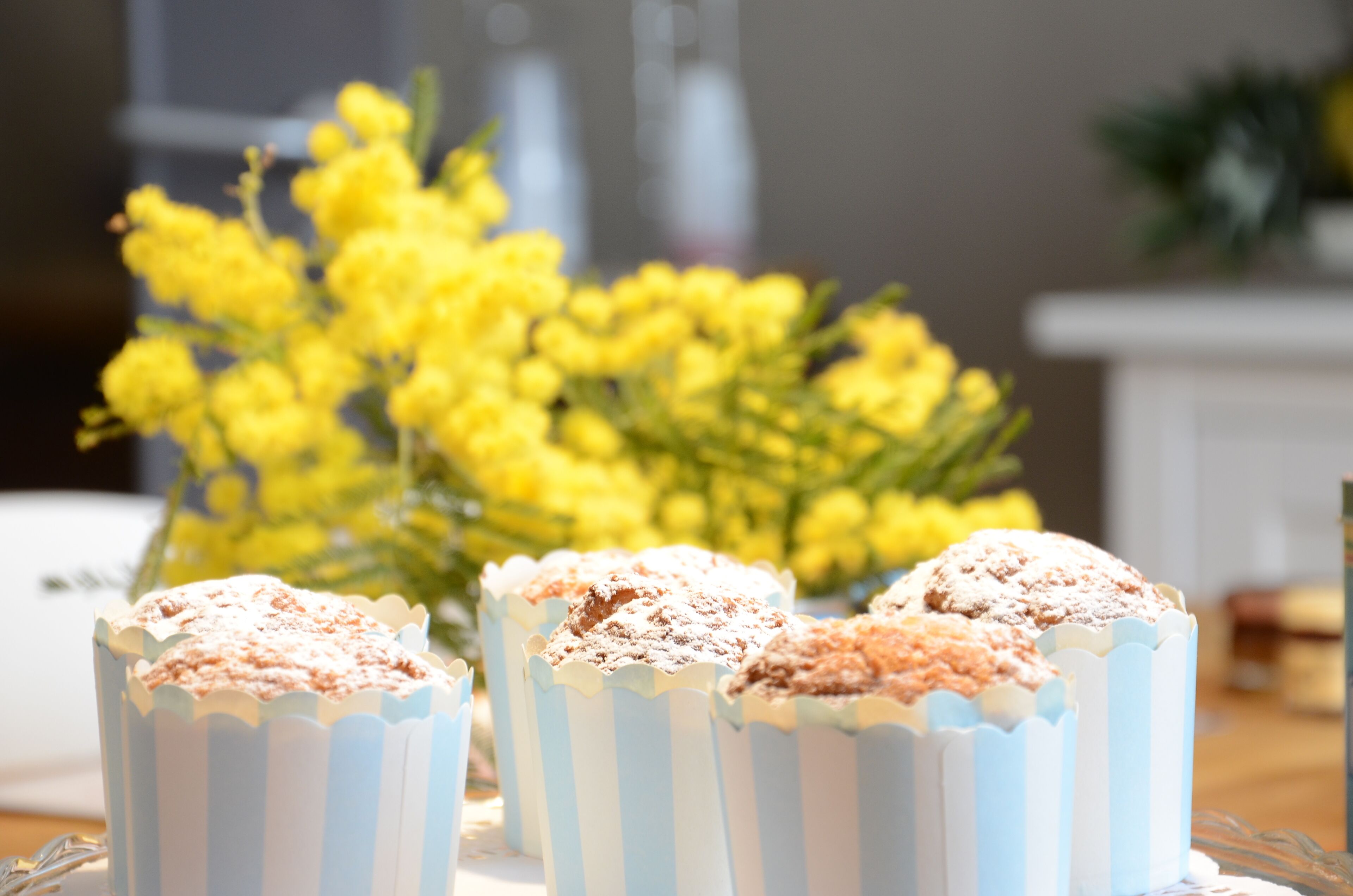 A table with cupcakes in striped paper cups, a vase of yellow flowers, and a white cabinet in the background.