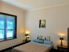 Un dormitorio con cama, dos mesitas de noche con lámparas, una ventana con persianas y un cuadro en la pared.
