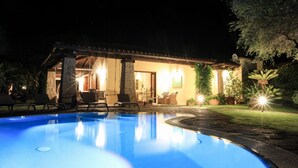 A nighttime view of a house with a swimming pool, outdoor seating, and a well-lit patio area.