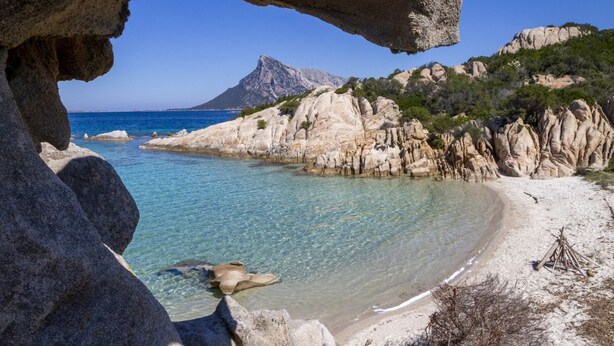A coastal beach with clear waters, rocky cliffs, and a mountain in the background.