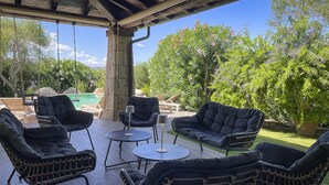 A covered outdoor seating area with black cushioned chairs and tables, a swing set, and a pool in the background.