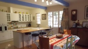 Een keuken met een centraal eiland, barstoelen, een eettafel met een tafelkleed met bloemenmotieven en een klok aan de muur.