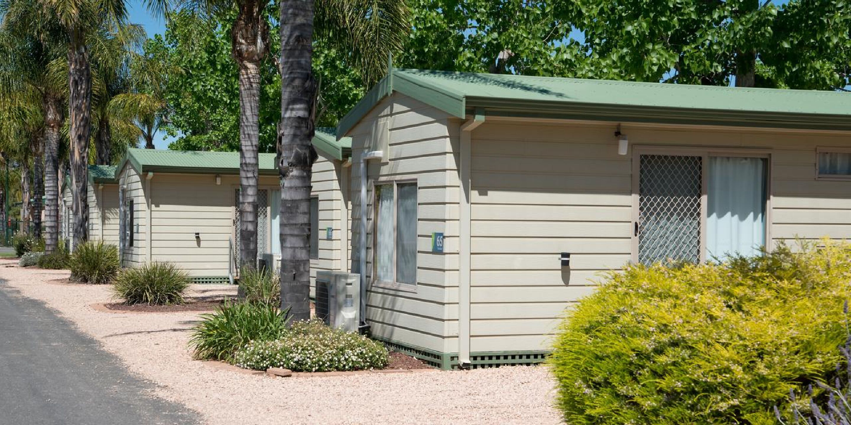 A row of single-story mobile homes with green roofs, surrounded by palm trees and small shrubs.