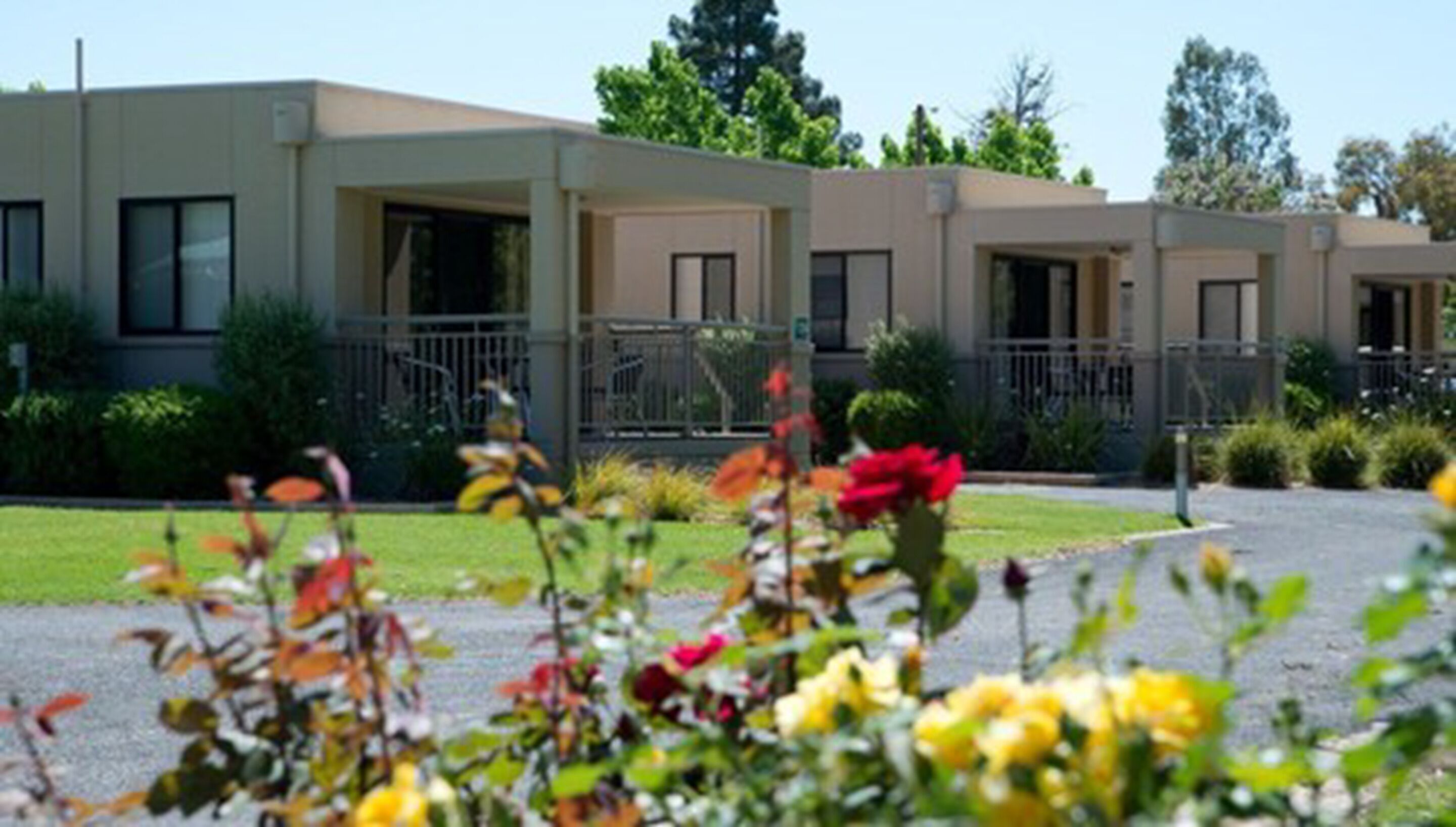 A row of modern, single-story buildings with balconies, surrounded by well-maintained lawns and flowering plants.