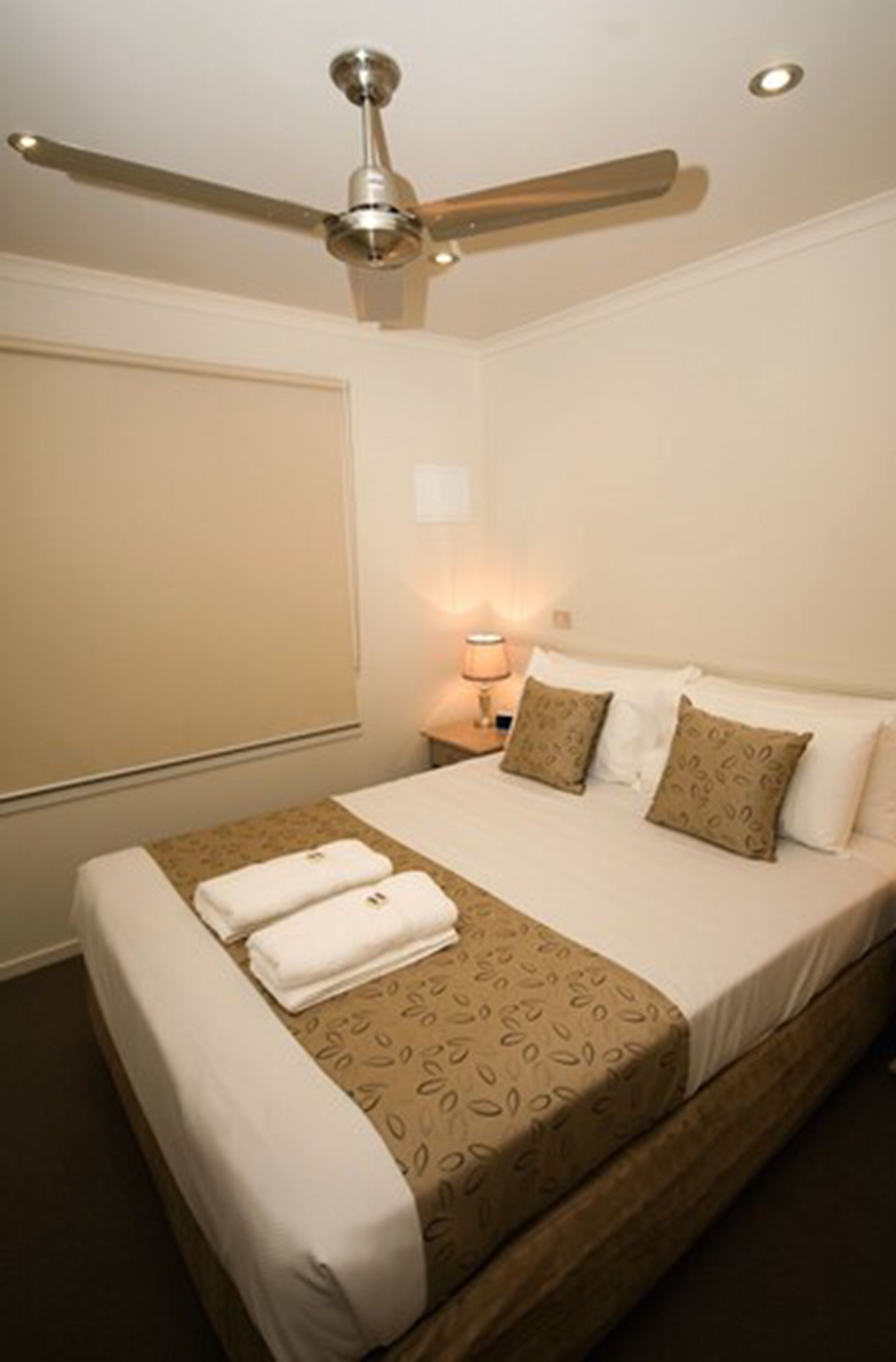 A hotel room with a bed, a ceiling fan, a nightstand with a lamp, and a window with blinds.