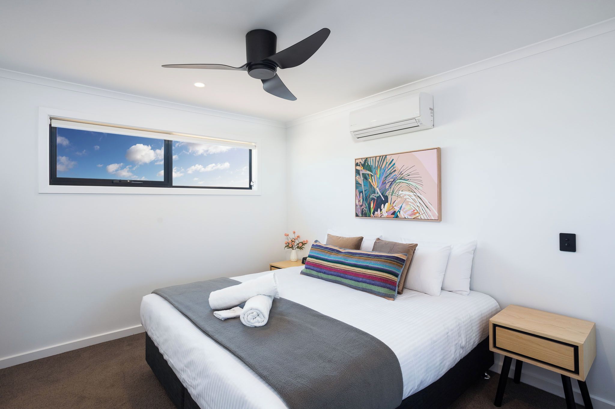Ein Schlafzimmer mit Bett, Deckenventilator, Klimaanlage, Fenster mit Blick auf den Himmel und einem farbenfrohen abstrakten Gemälde an der Wand.
