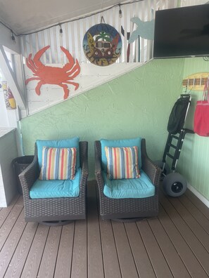 A covered outdoor seating area with wicker chairs and striped cushions, a wall with surf-themed decorations, and a wall-mounted TV.