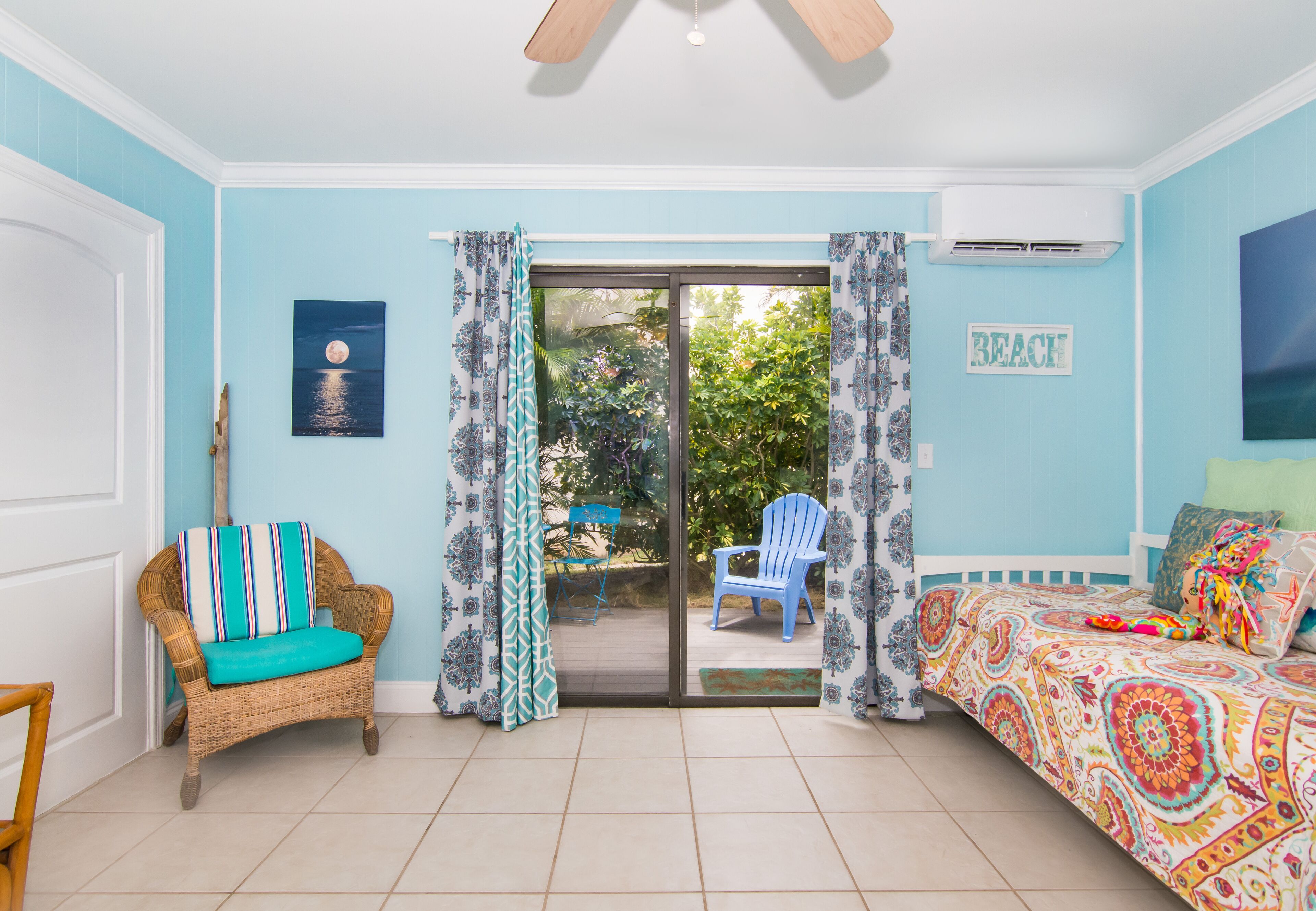 Una habitación con una silla de mimbre, una cama colorida y vistas a un área exterior con una silla y plantas a través de un ventanal corredero.