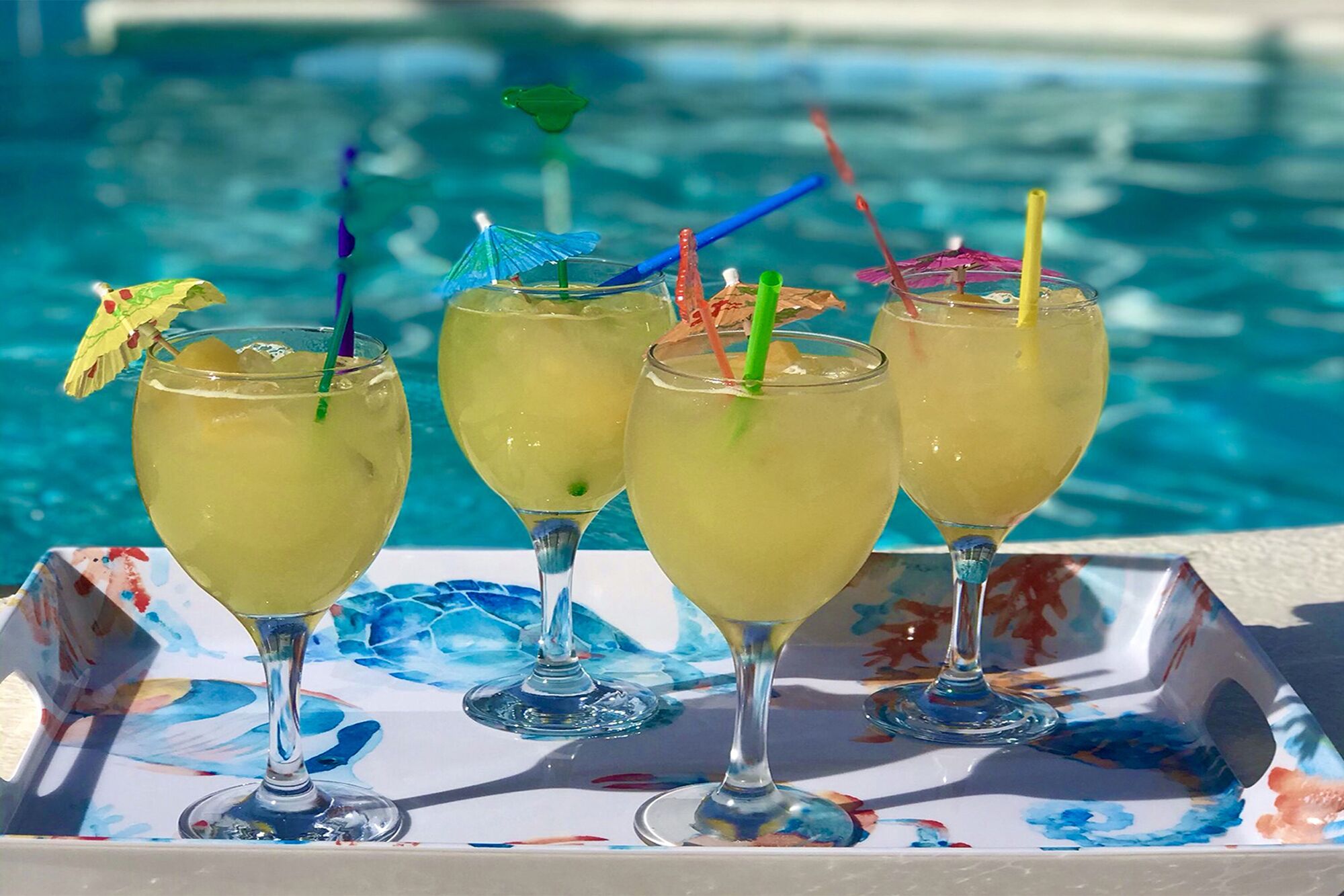 Four glasses of a yellow beverage with colorful straws and umbrellas, placed on a tray by a pool.