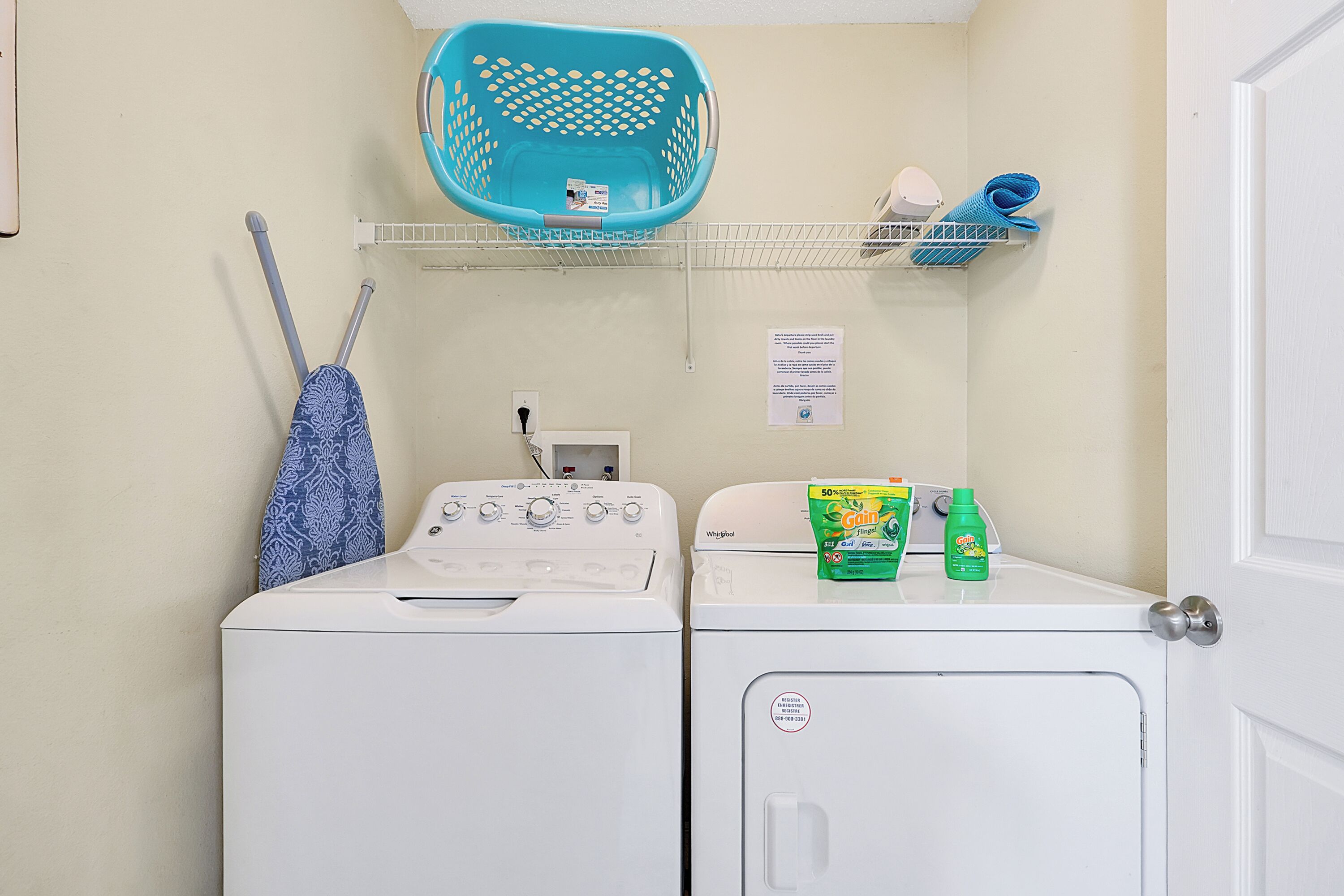 A laundry area with two washing machines, a blue laundry basket, and a shelf with cleaning supplies.