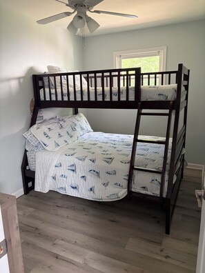 A bunk bed with a ceiling fan, a window with a view of greenery, and a wooden floor.