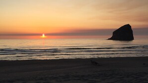 A beach with a large rock formation and the sun setting over the ocean.