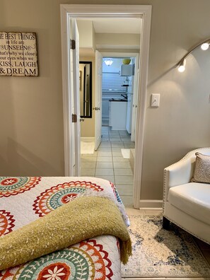Un dormitorio con cama, una silla, un adorno en la pared y vistas a la cocina.
