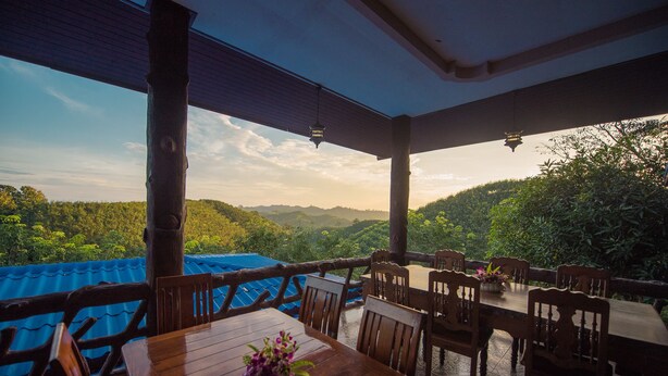 A wooden table and chairs set up on a terrace with a view of a lush green landscape and hills in the distance. - Mountain View House (Sangkhla Buri)