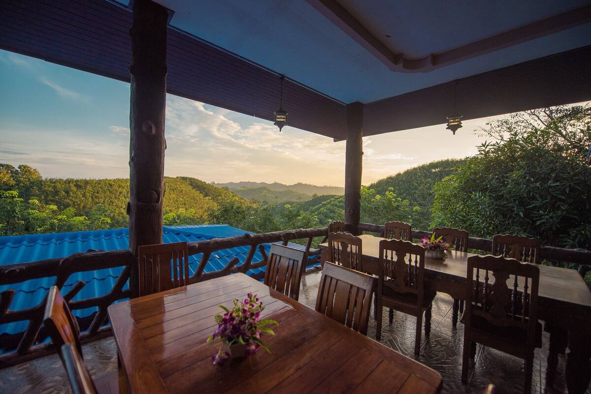 A wooden table and chairs set up on a terrace with a view of a lush green landscape and hills in the distance.