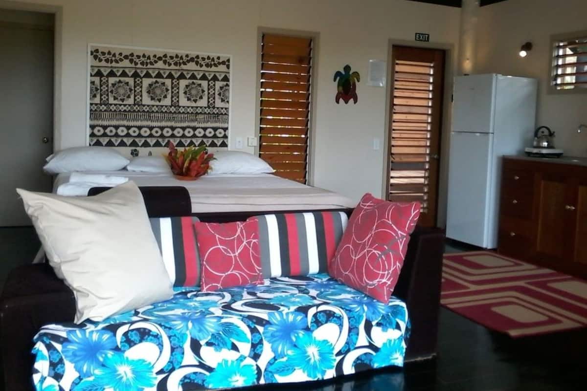 Un dormitorio con cama, sofá, una pared con diseño decorativo, refrigerador y ventana con persianas.