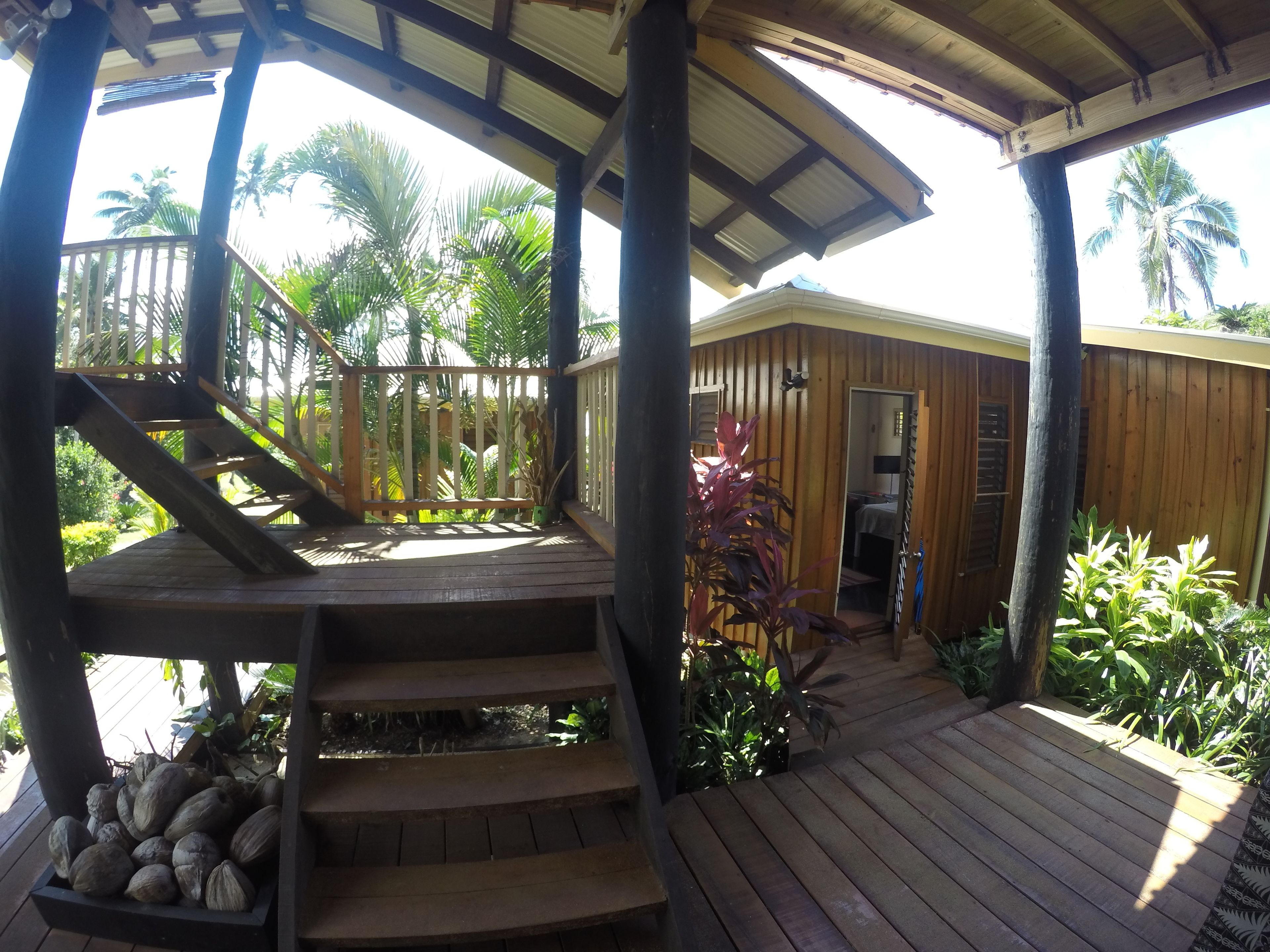 A wooden deck with a staircase leading to a covered area, surrounded by lush greenery and palm trees.
