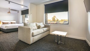 Ein Hotelzimmer mit einem Bett, einer Couch, einem kleinen Tisch und einem großen Fenster mit Blick auf Autos und Bäume.