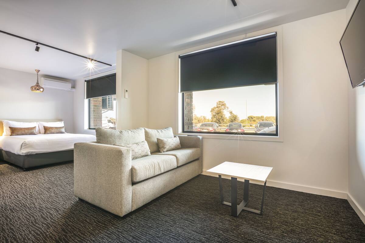 A hotel room with a bed, a sofa, a small table, and a large window with a view of cars and trees.