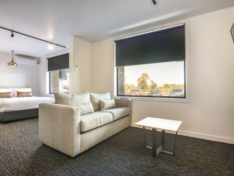 Habitación de hotel con cama, sofá, una mesita y un ventanal con vistas a coches y árboles.
