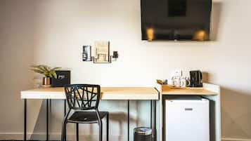 Een compacte hotelkamer met een eenpersoonsbed, een bureau met stoel, een kleine koelkast, een aan de muur bevestigde televisie en een potplant.