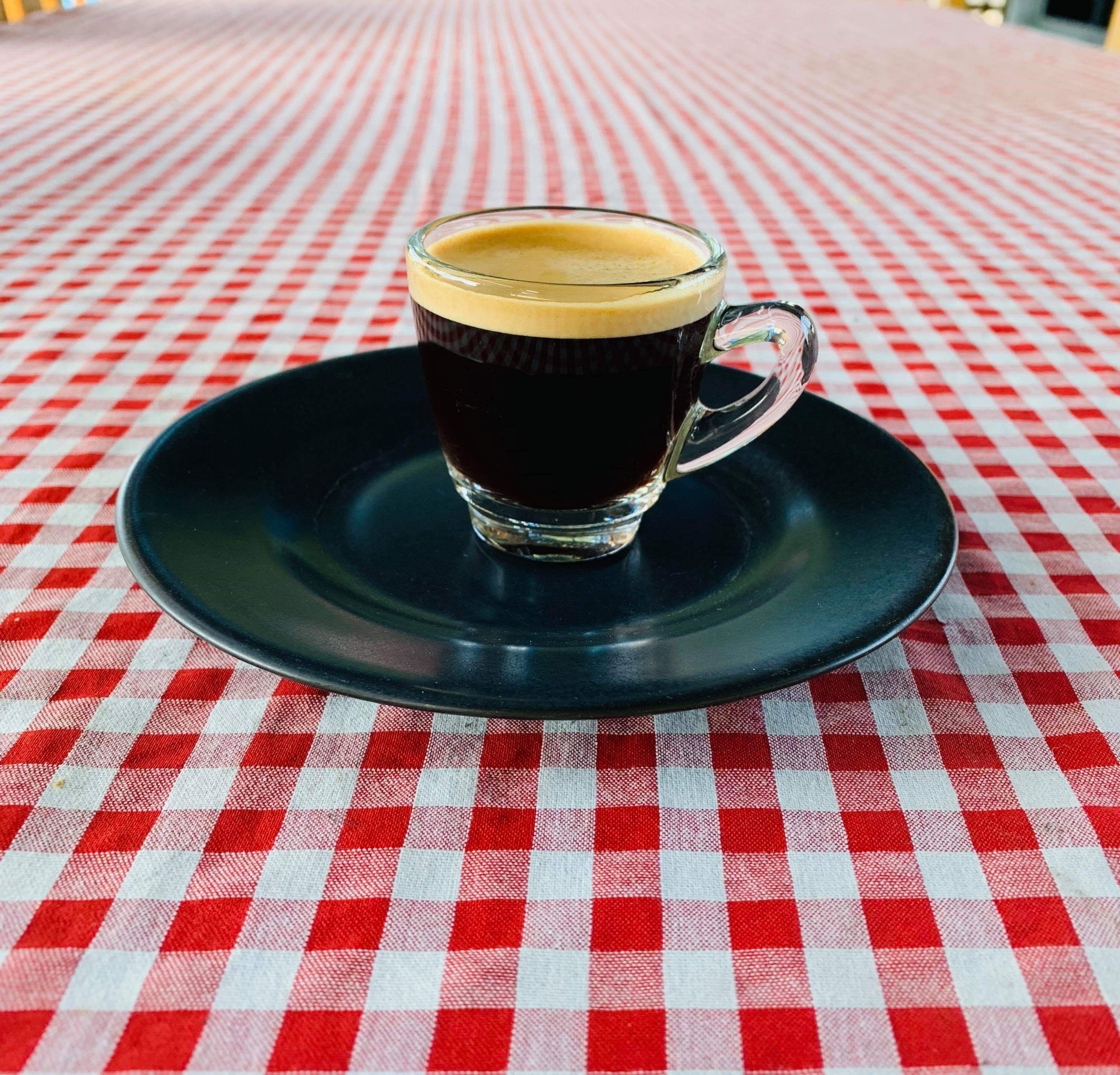 A glass cup of dark coffee on a black saucer, placed on a red and white checkered tablecloth.