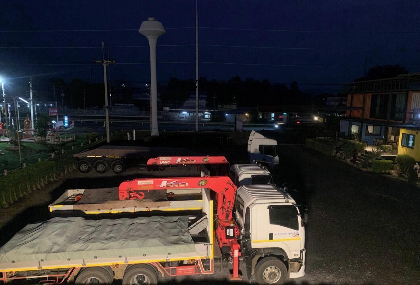Cena noturna com vários camiões estacionados na estrada, um poste de luz alto e edifícios ao fundo.