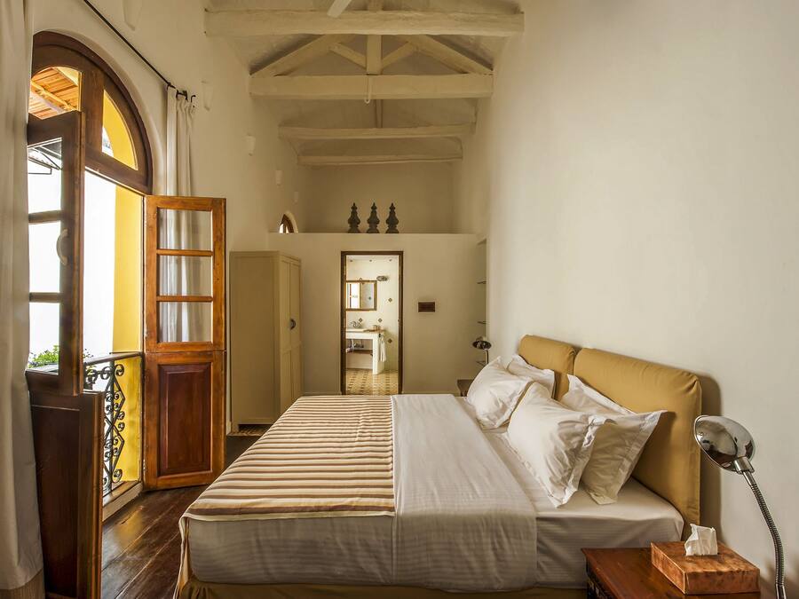 Un dormitorio con cama, mesita de noche, espejo y vista a un baño a través de una puerta abierta.
