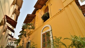 A narrow alleyway with yellow buildings, a visible window with a wooden frame, and potted plants along the path. - Mateus Boutique Hotel (Panaji)