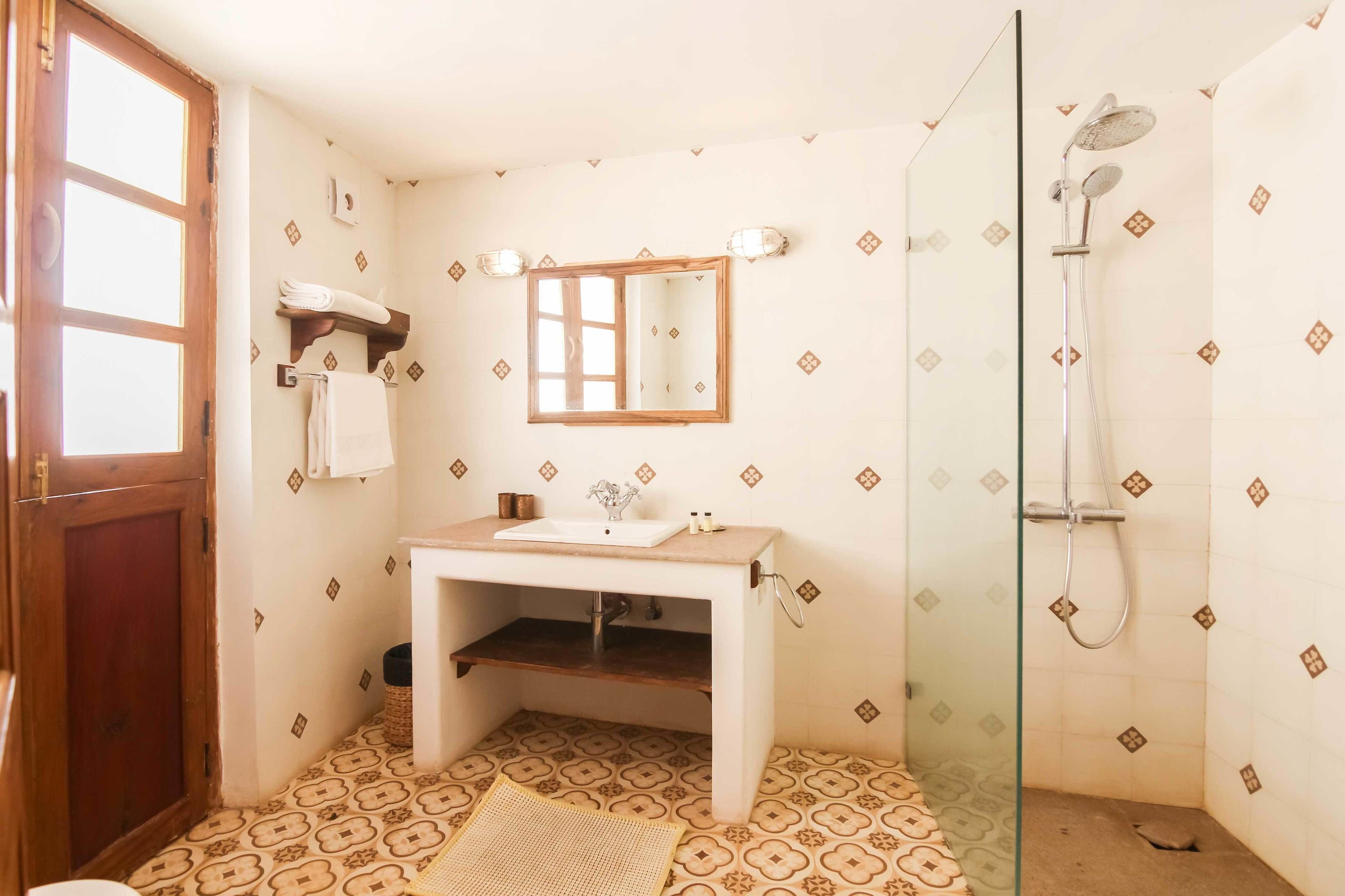 A bathroom with a wooden door, a white sink with a mirror above it, a shower with a glass door, and patterned floor tiles.