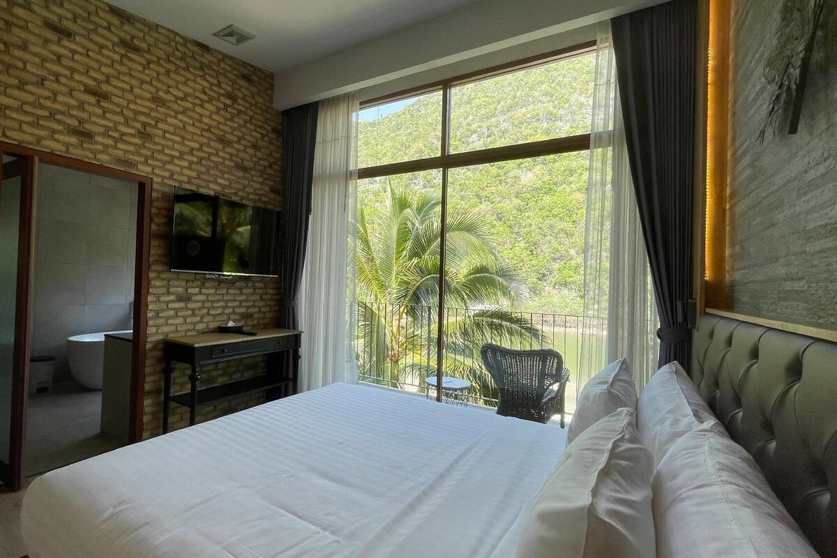 Een hotelkamer met een bakstenen muur, een groot bed met witte lakens, een bureau met een flatscreen televisie en een balkon met een stoel en tafel.