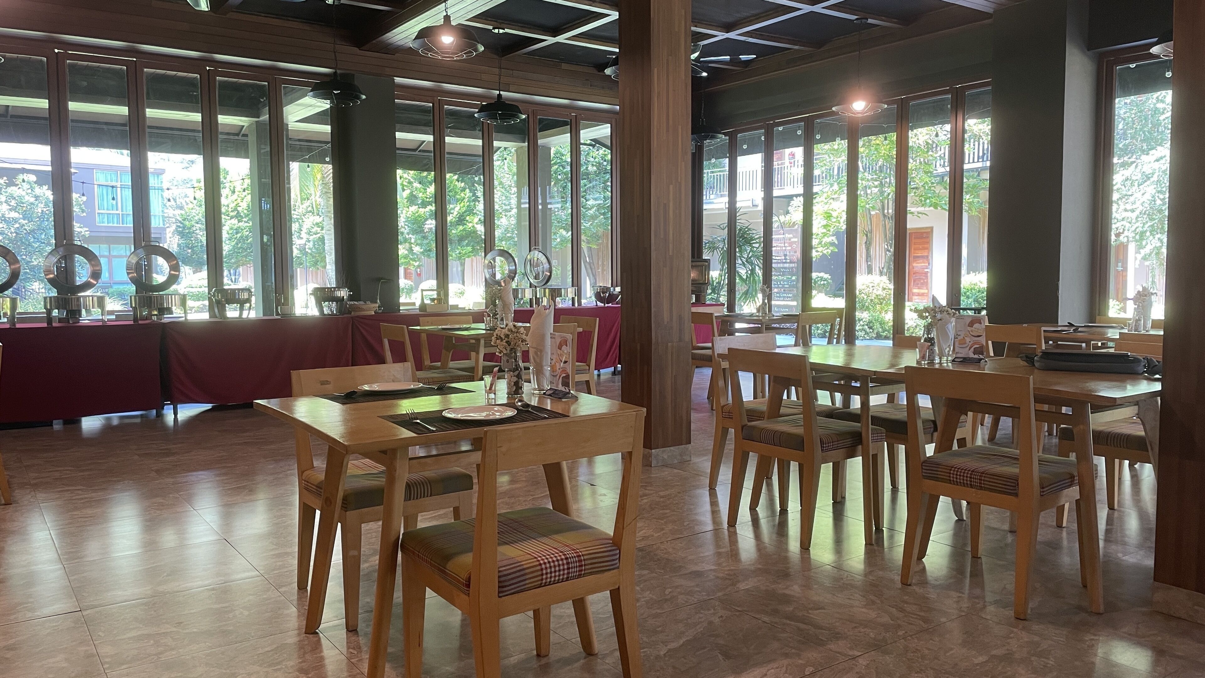 A spacious restaurant with wooden tables and chairs, large windows offering views of greenery outside.