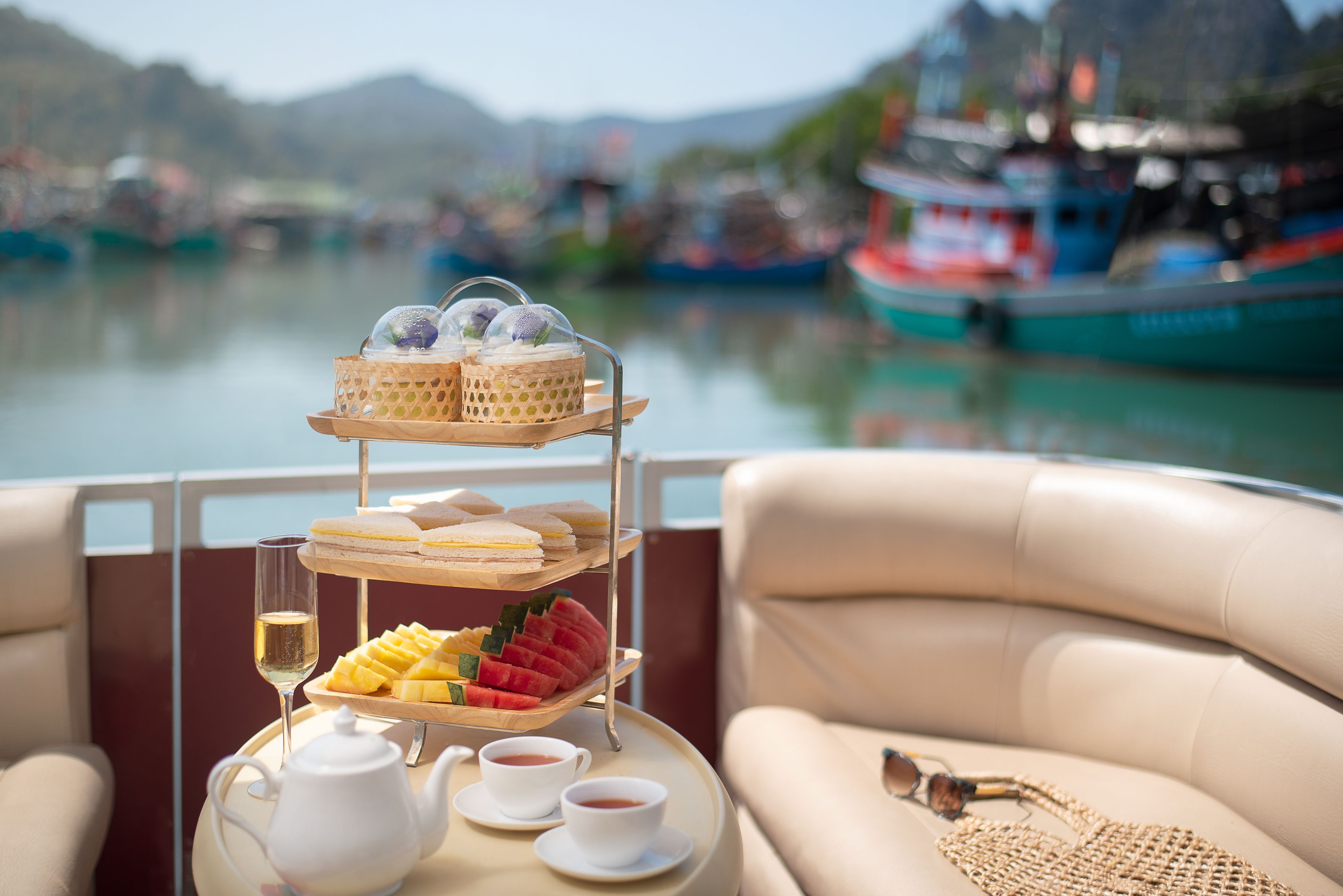 A tiered tray with sandwiches, fruit, and tea on a boat with a view of boats and mountains.