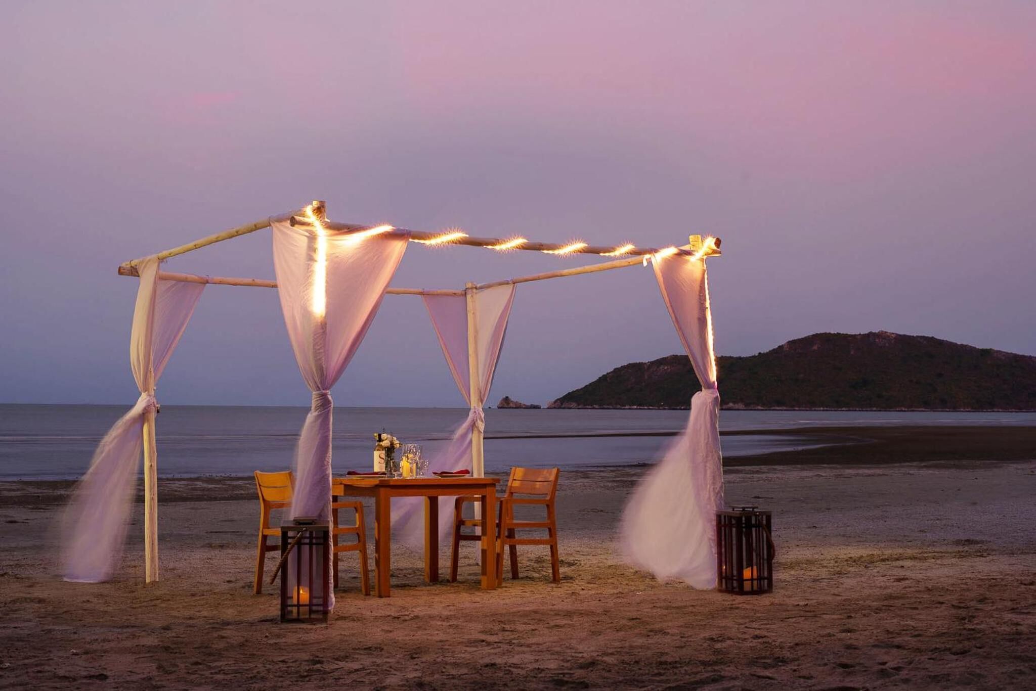 Un montaje para comer frente al mar, con una mesa y sillas bajo una estructura cubierta, rodeado de faroles y telas decorativas.