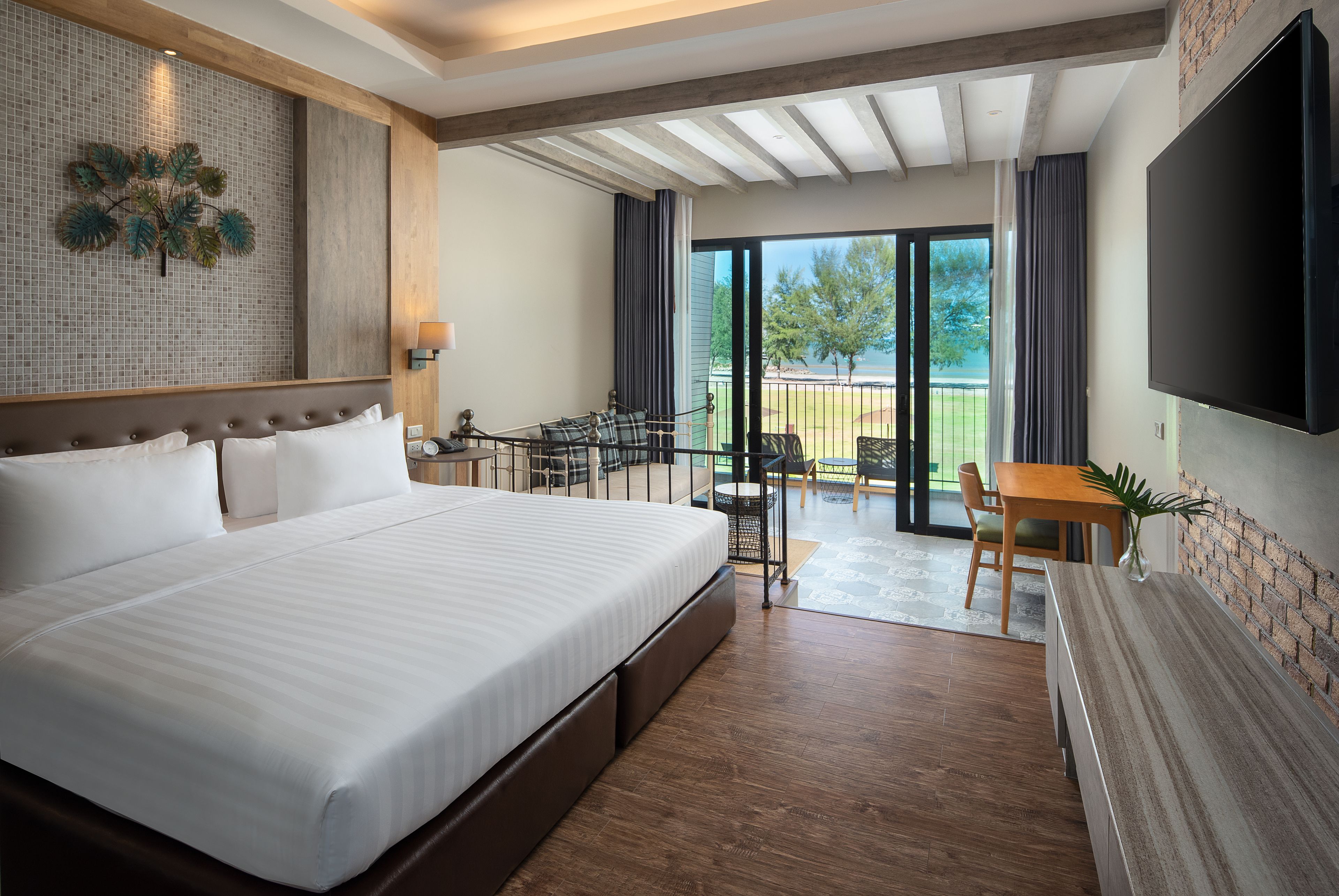 A modern hotel room with a large bed, a dining area with a table and chairs, a TV mounted on the wall, and a balcony with a view of the outdoors.