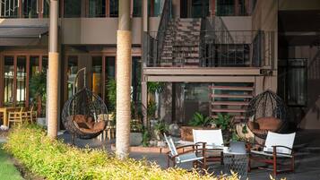 An outdoor seating area with wicker chairs and a hanging chair, surrounded by greenery and a building with a balcony.