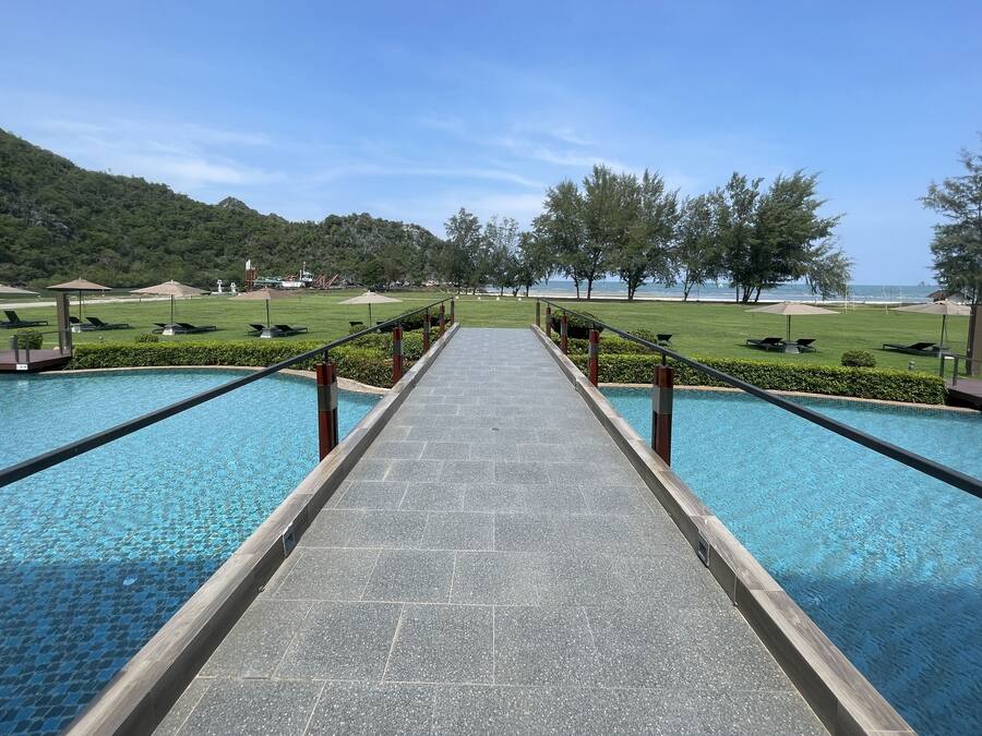Un espace piscine avec un chemin central menant à deux piscines, entouré de chaises longues et de parasols, offrant une vue sur une plage et des collines en arrière-plan.