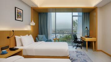 A hotel room with a bed, bedside table, lamp, desk, chair, and a view of the city.