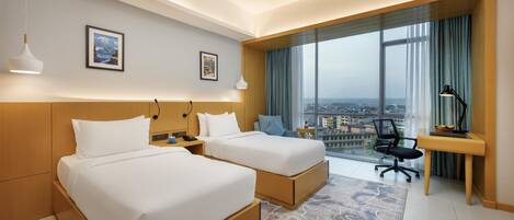 A hotel room with two beds, a desk, a chair, and a large window with a city view.