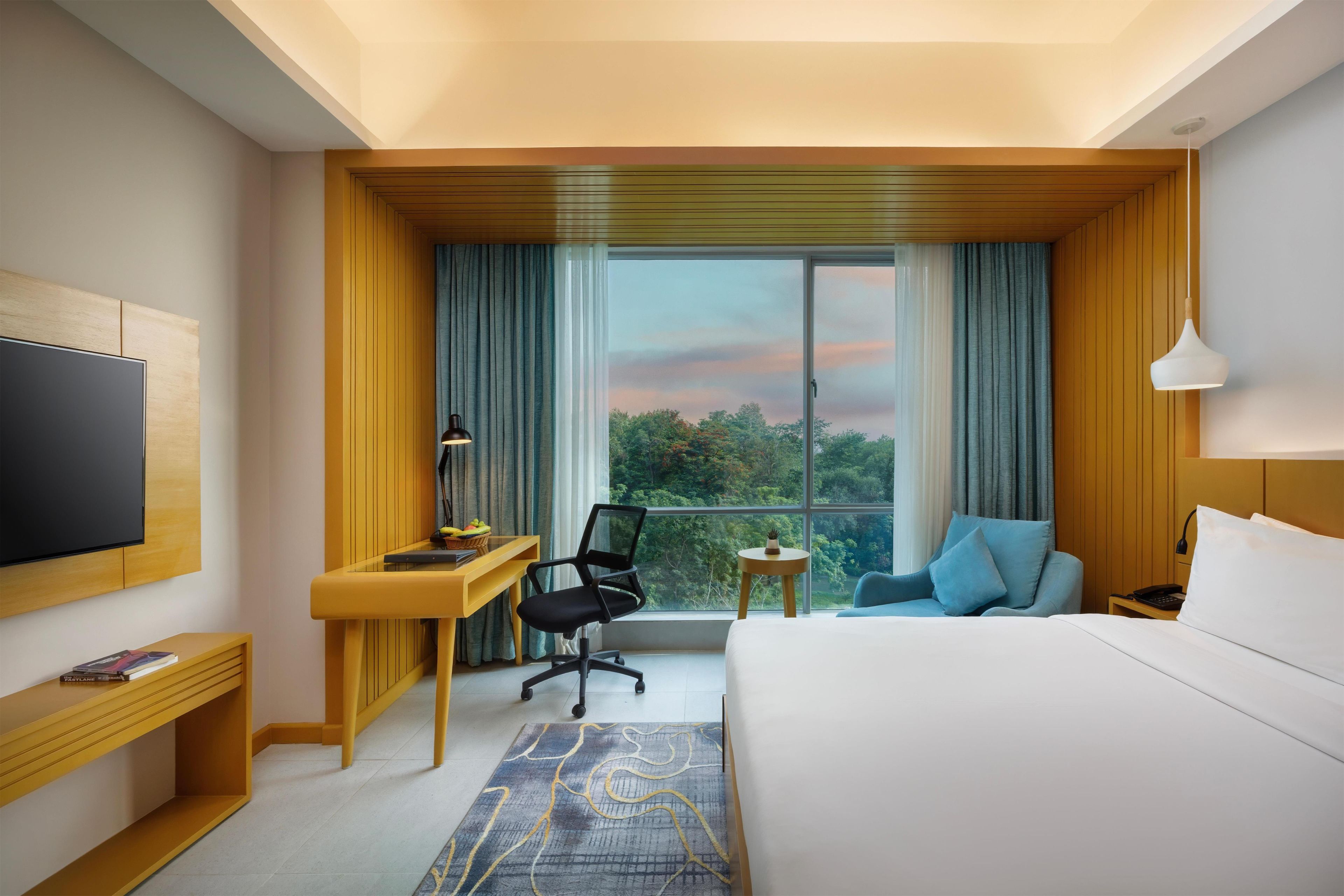 Habitación de hotel con cama, escritorio, silla, televisor y vista al exterior.
