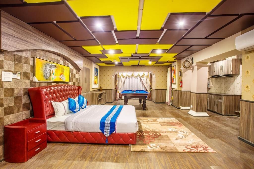 A hotel room with a red bed, a pool table, a kitchen area, and a clock on the wall.